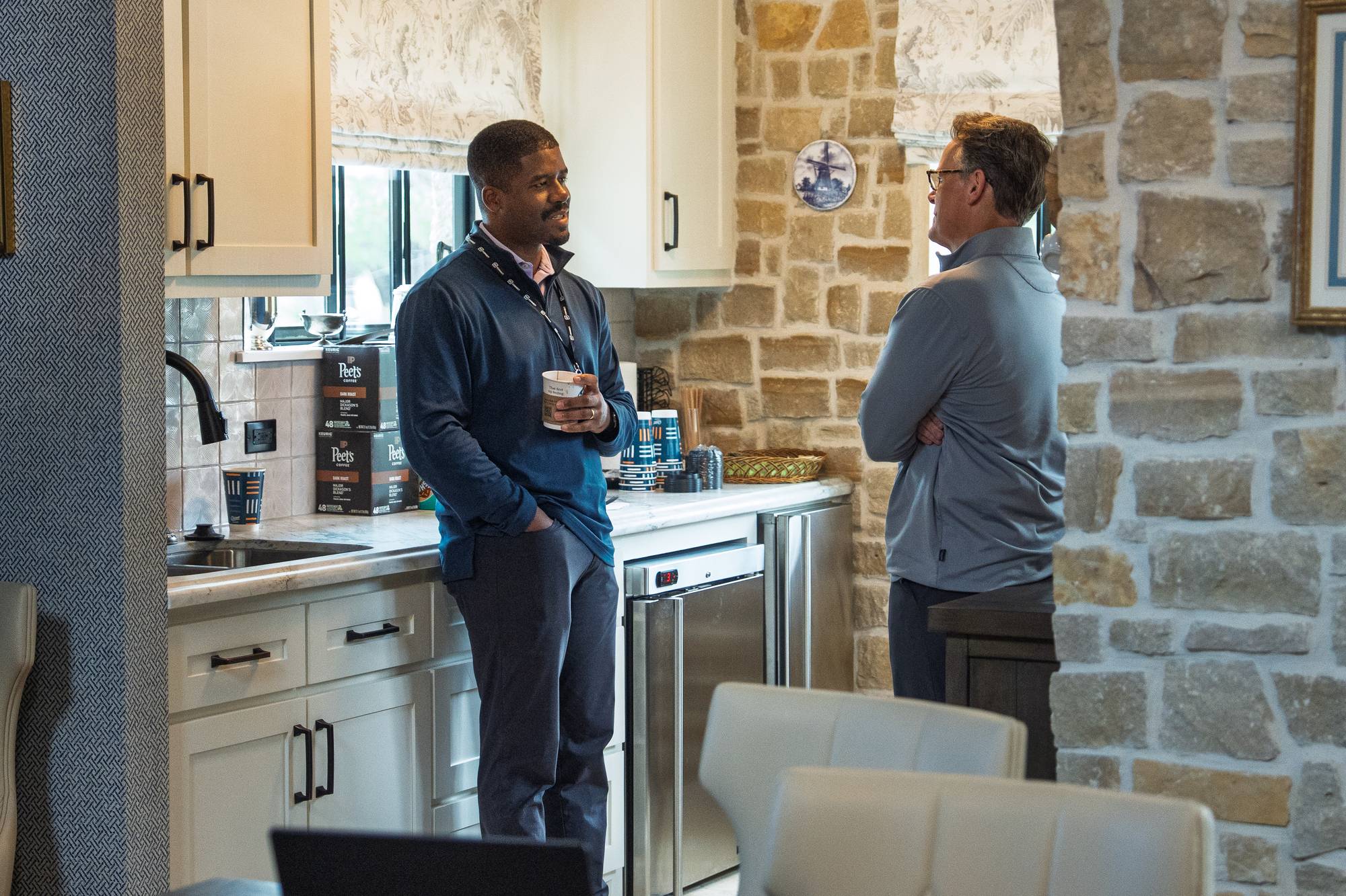 Steven Smith, arms crossed, listening as a pastor holding a coffee cup talks with him in a kitchen with stone walls