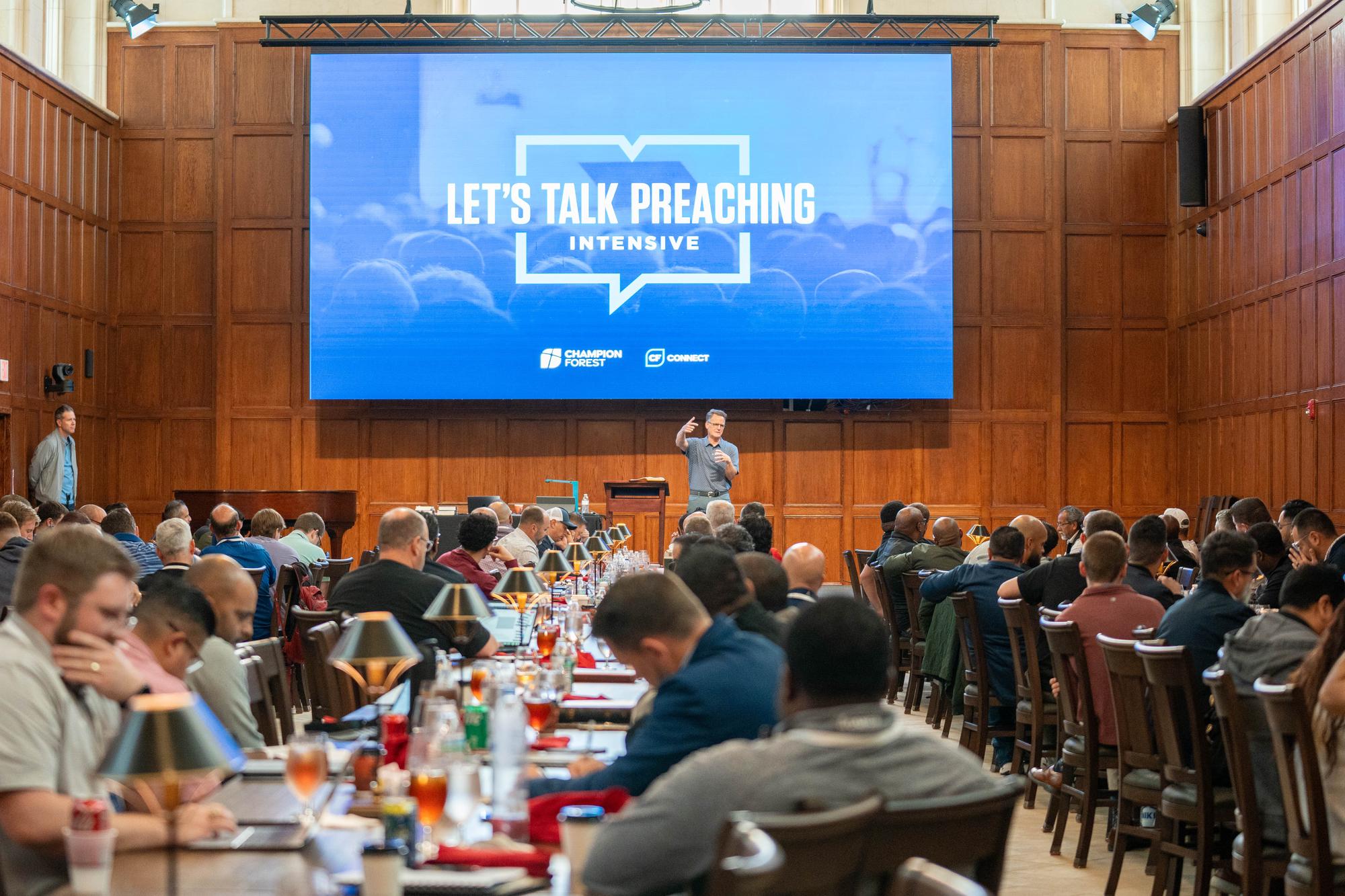 A wood-paneled library hall full of pastors at long tables with brass lamps, listening as Steven Smith teaches beneath a large screen