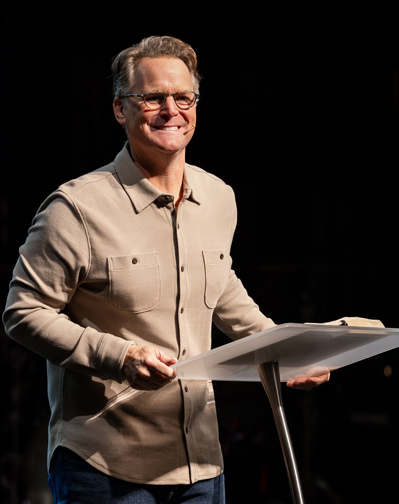 Steven Smith smiling behind a clear lectern on a dark stage, wearing a tan button-up shirt
