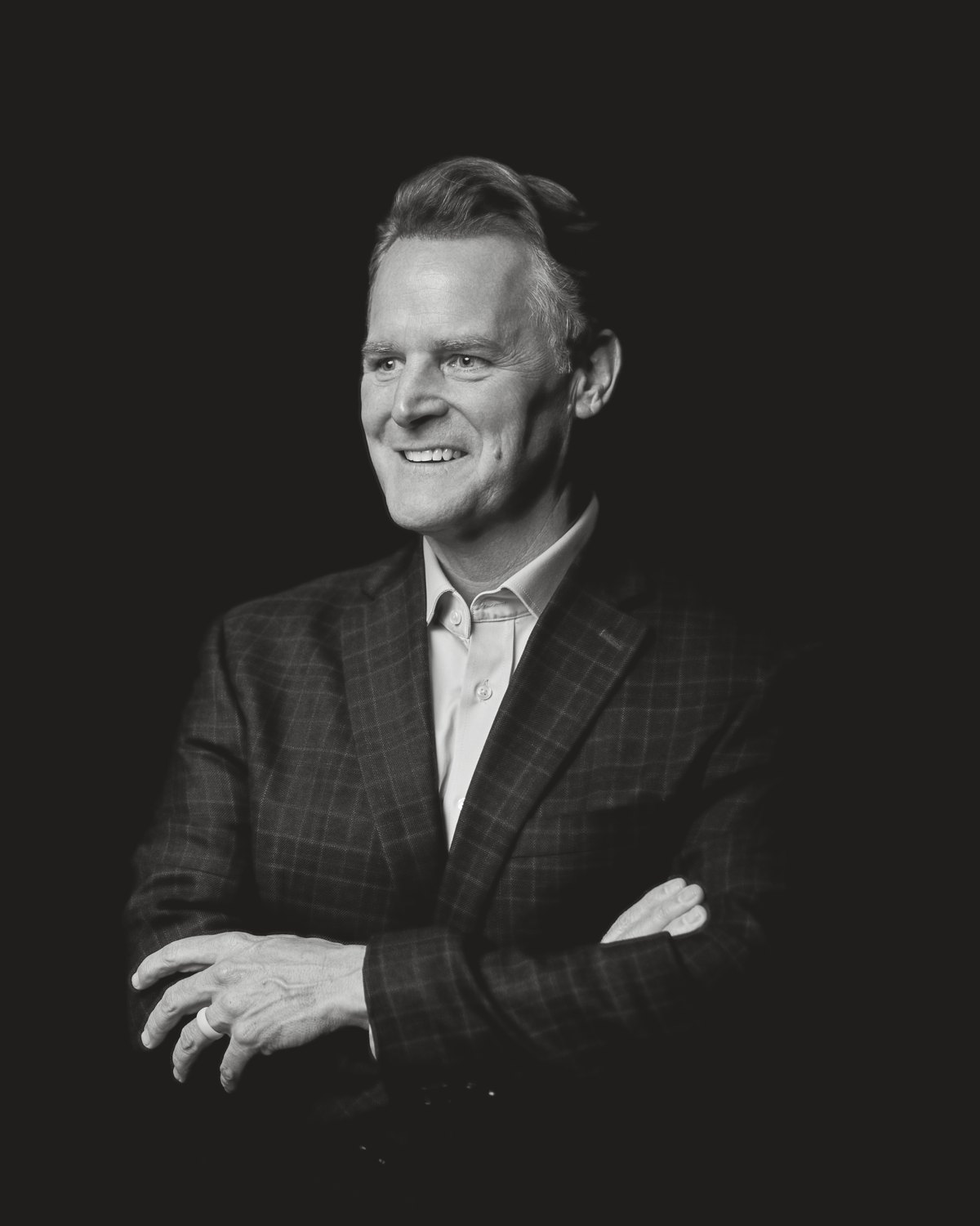 Black-and-white portrait of Steven Smith smiling, arms crossed, in a checked sport coat and open-collar shirt against a dark backdrop